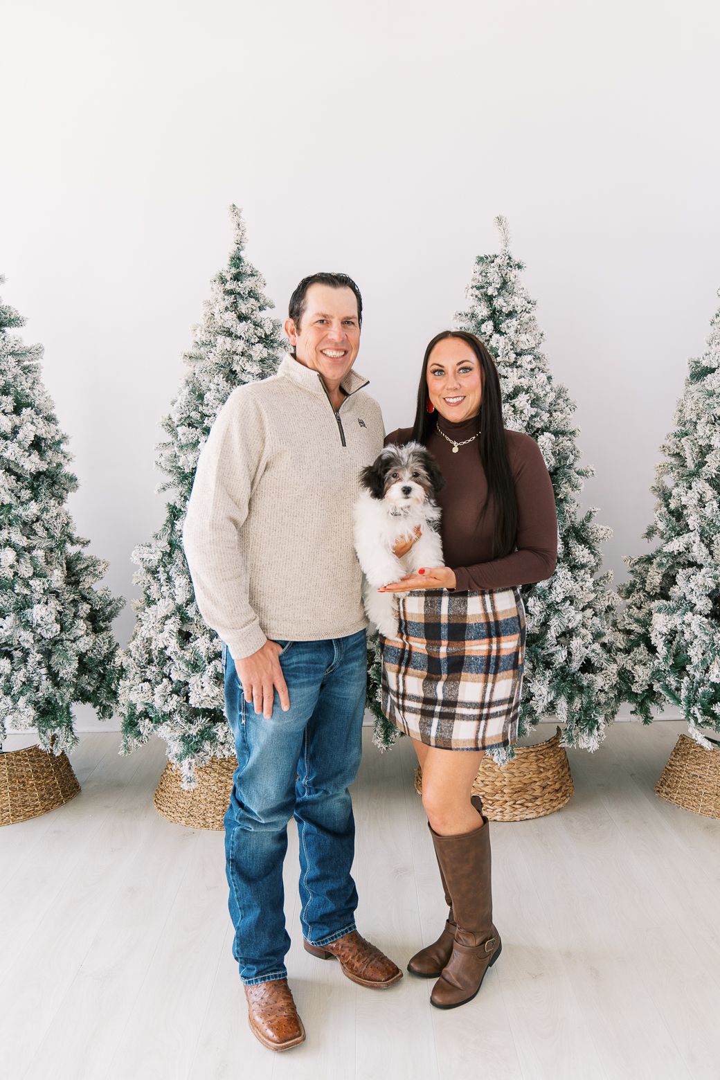 A couple poses with their small dog in front of snowy Christmas trees and wicker baskets in a winter photo setting.