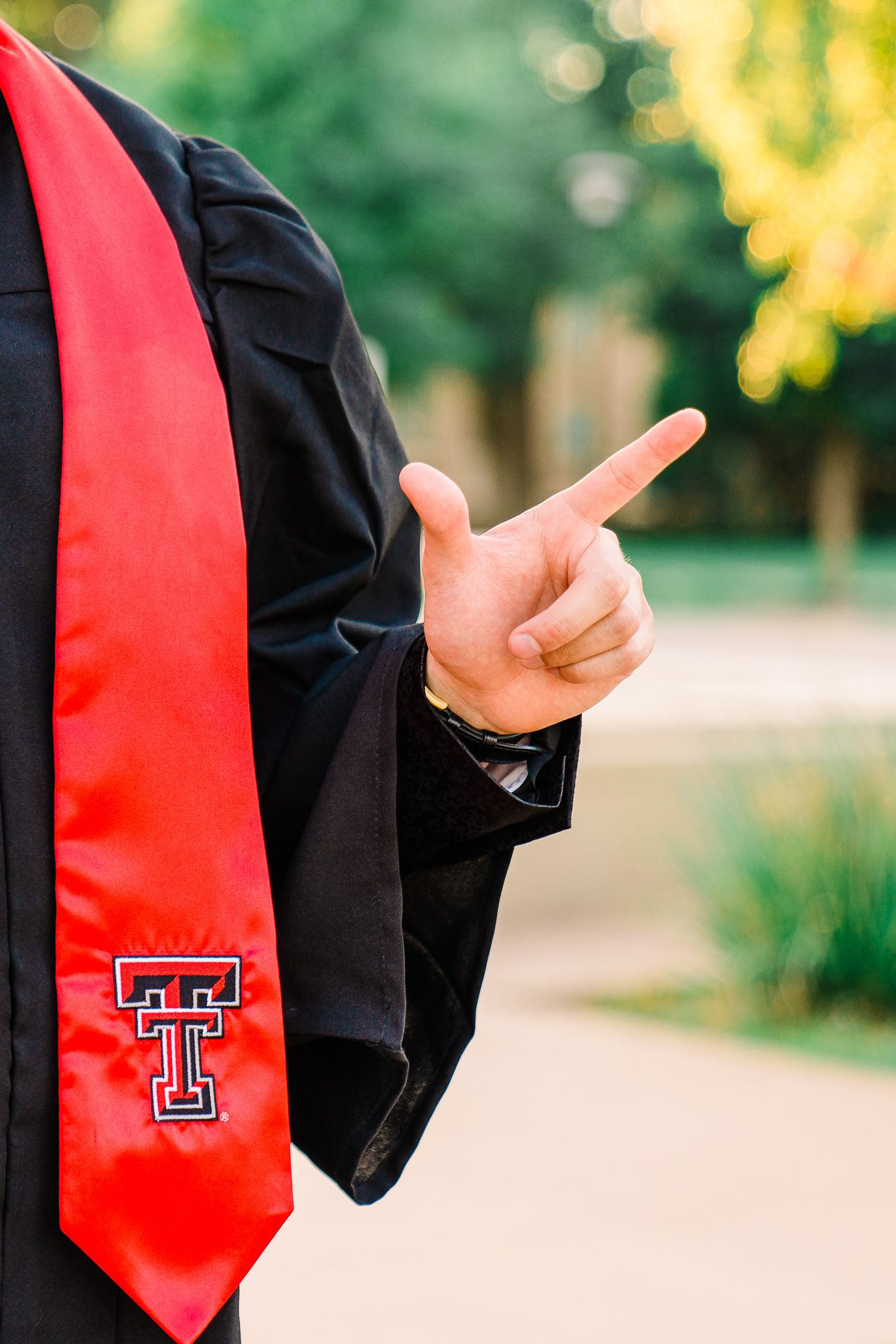 Graduate wearing black gown and red Texas Tech stole points finger while posing outdoors with blurred background.