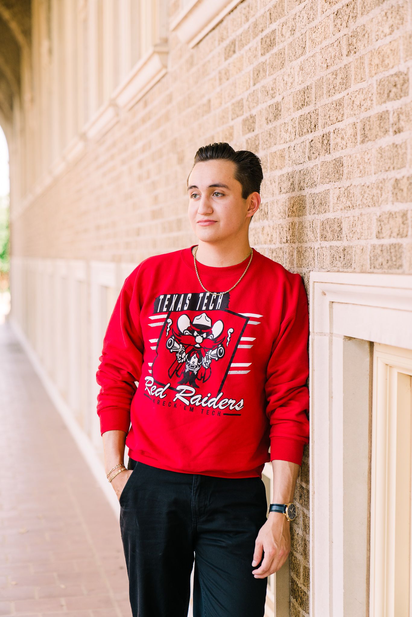 A person wearing a red Texas Tech Raiders sweatshirt and black pants leans against a beige brick wall.
