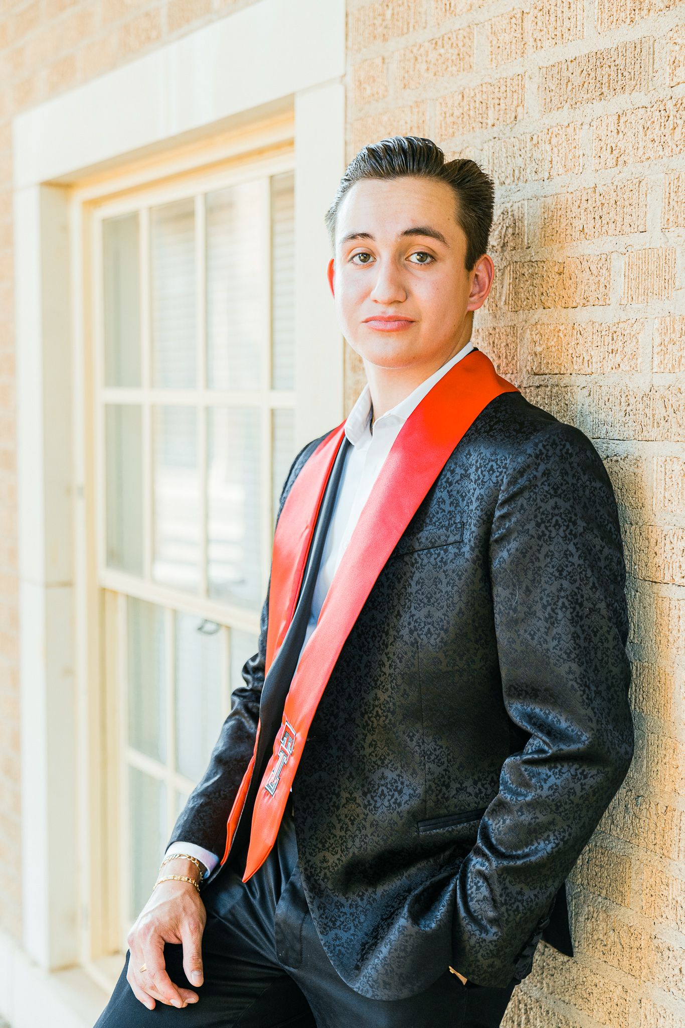 Graduate in dark suit and red stole poses against brick wall with windows.