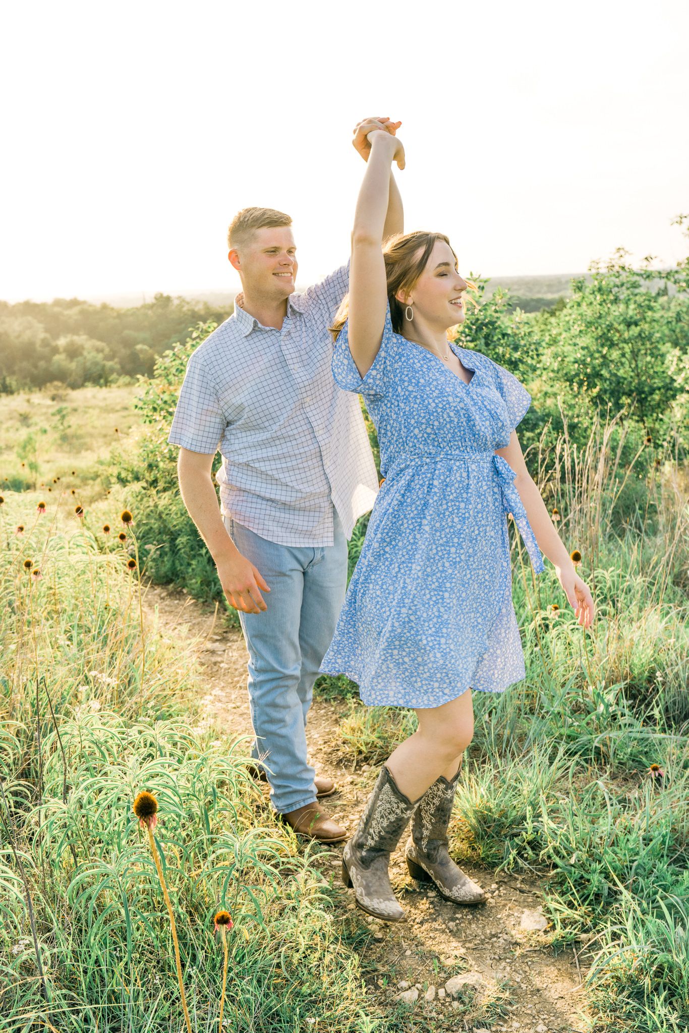 A couple walks hand in hand through a sunlit field with wildflowers and greenery during a romantic outdoor photoshoot.