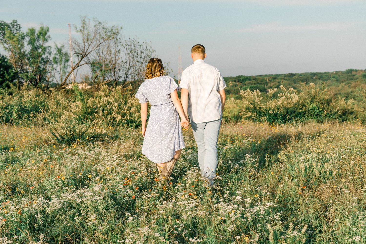 A couple in white outfits walks together through a sunny wildflower meadow on a summer afternoon.