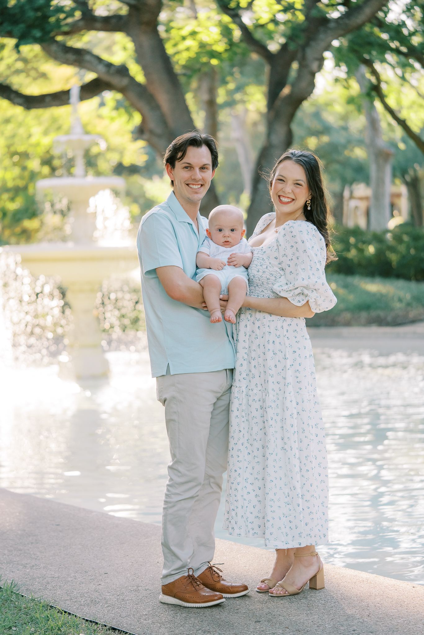 Happy young family with a baby posing by a fountain in a lush park setting.
