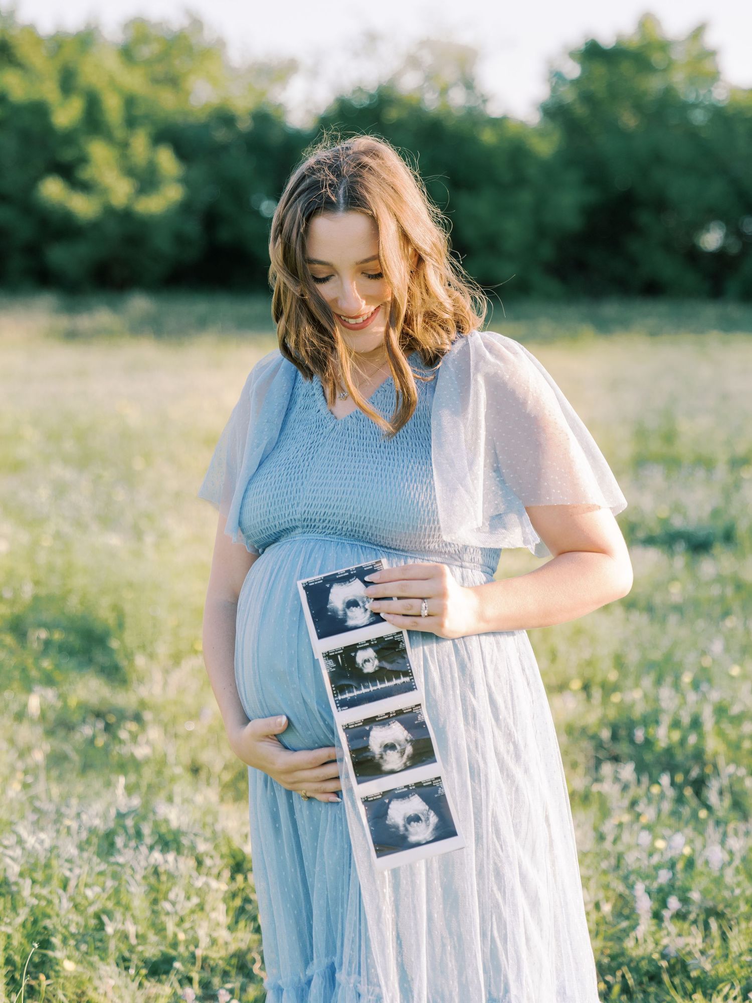 Pregnant woman in blue dress holding ultrasound photos in a flower field during golden hour maternity photoshoot.