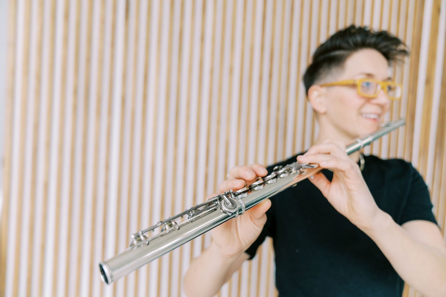 Musician in black t-shirt plays silver flute against striped background in warm lighting.