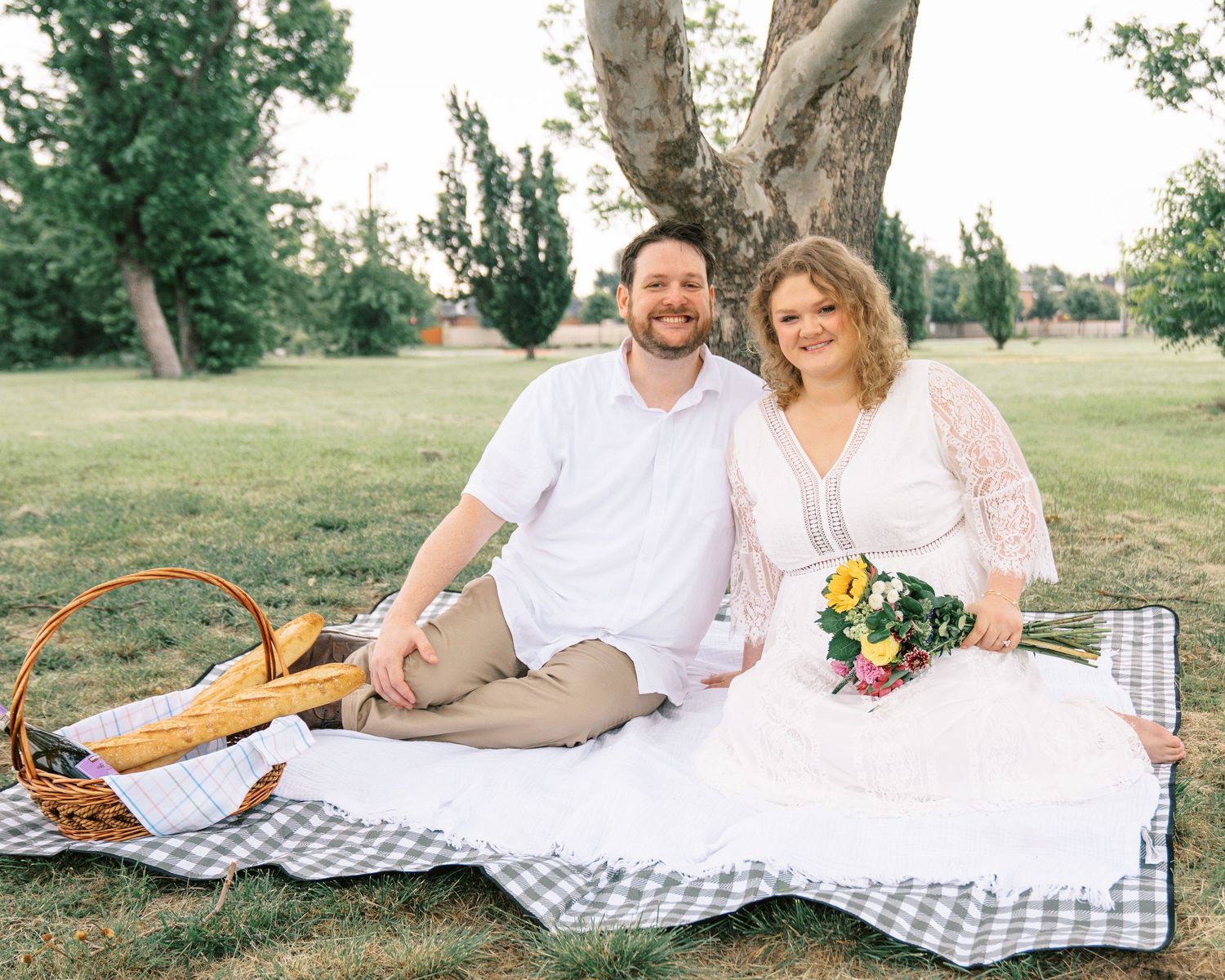 Couple in white wedding attire sitting on a picnic blanket under a tree with a wicker basket.