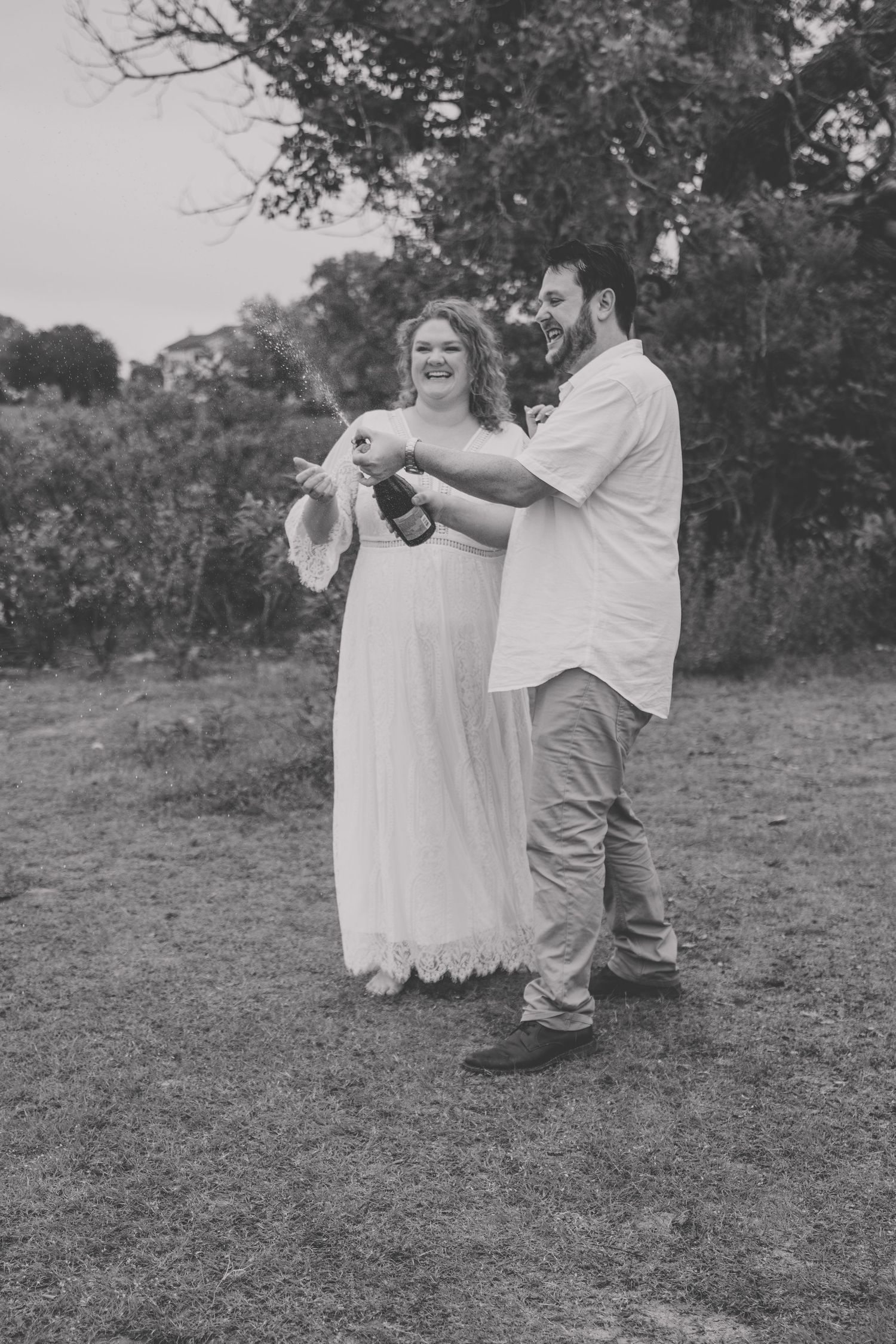 Black and white photo of a couple opening a champagne bottle outdoors on grass with trees in the background.