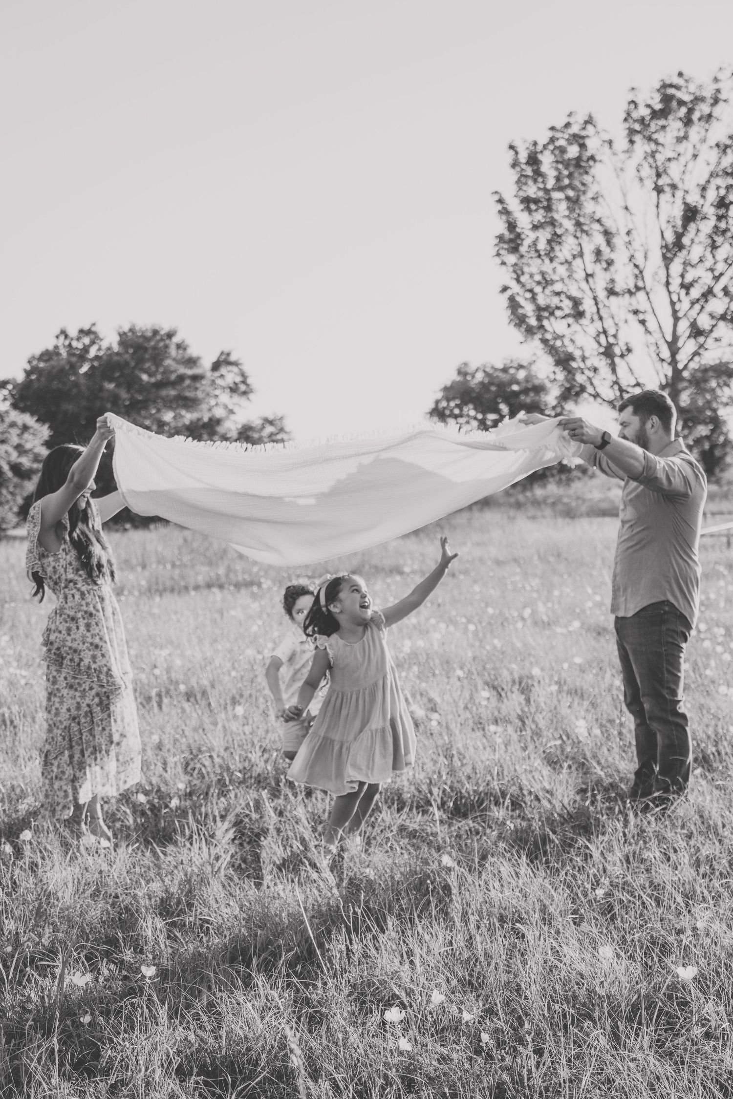 Black and white photo of a family playing with a blanket in a sunny meadow, with trees in the background.