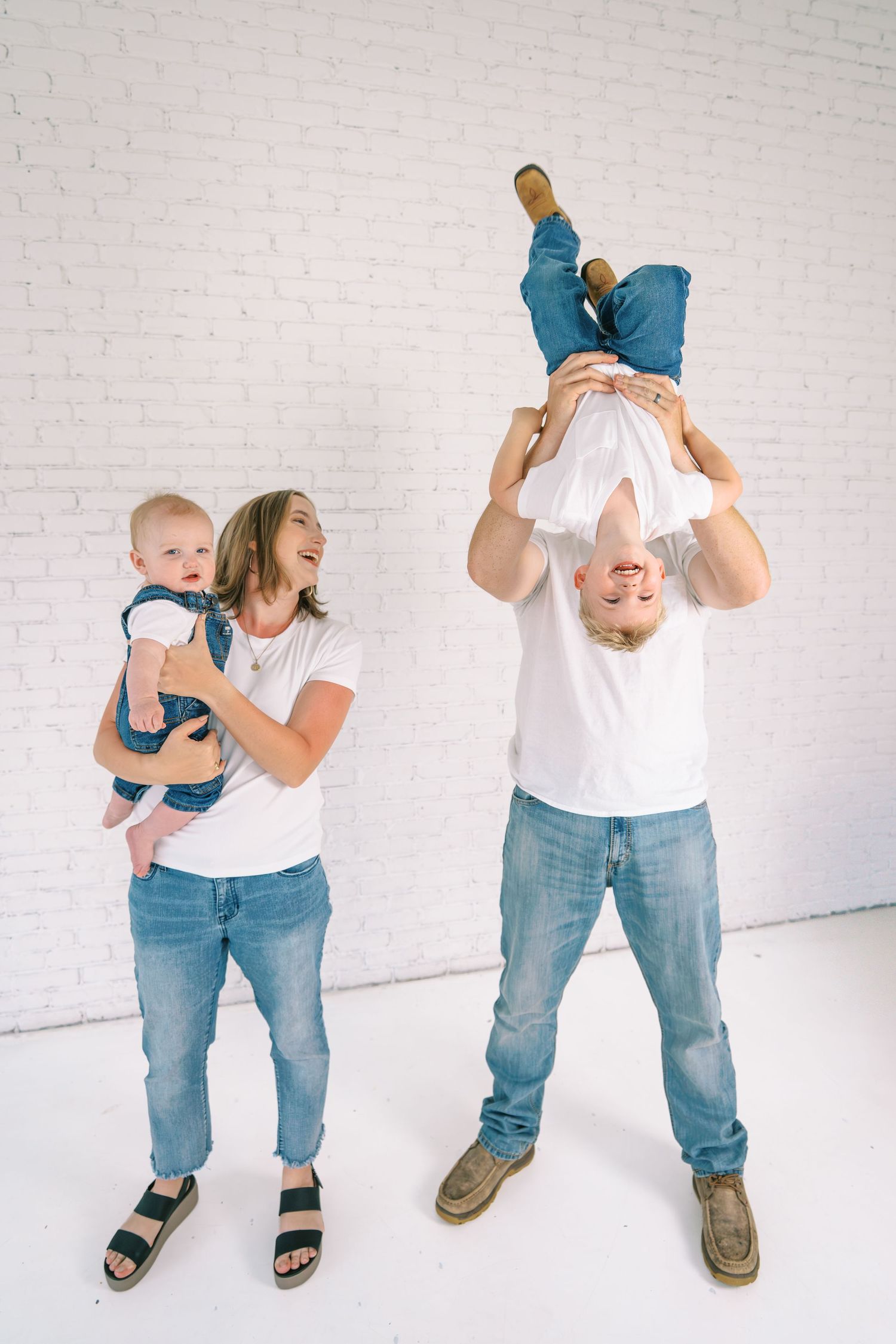 Family in white tees and jeans, mom holds baby while dad playfully lifts toddler upside down against white brick wall.