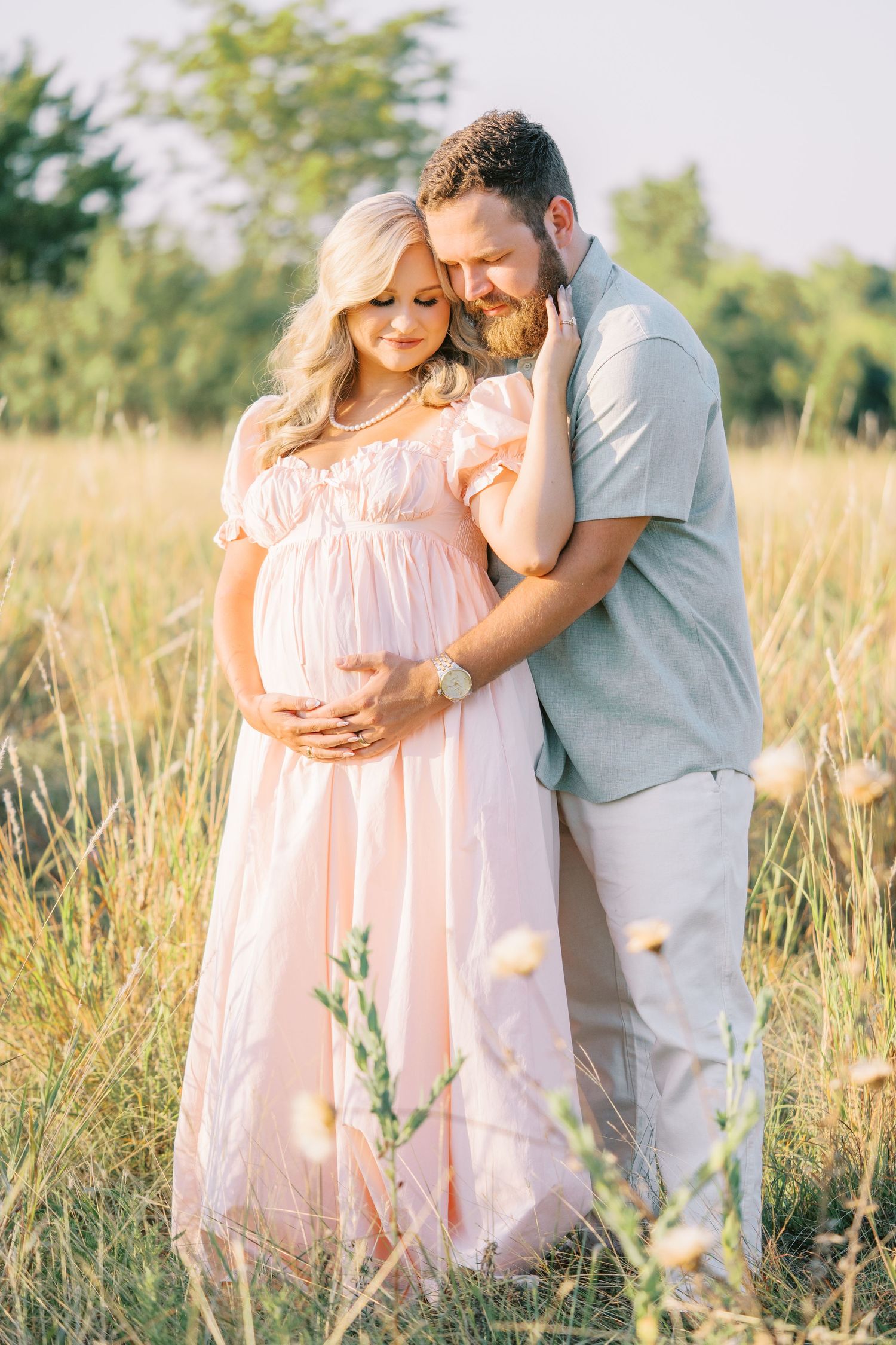Pregnant woman in pink dress embraced by man in field during golden hour maternity photoshoot.
