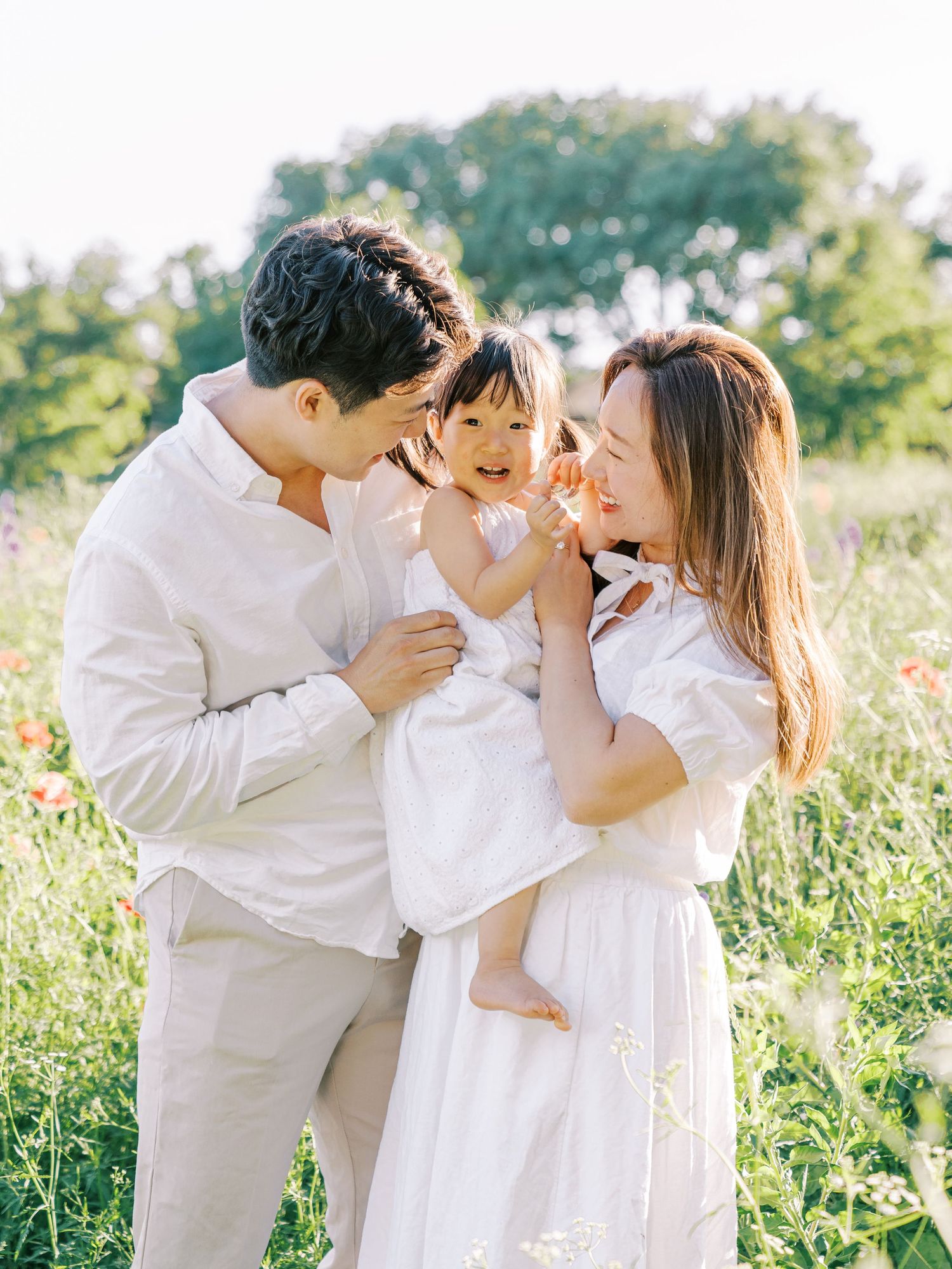 Family wearing white outfits shares tender moments together in a sunlit meadow during outdoor photo session.