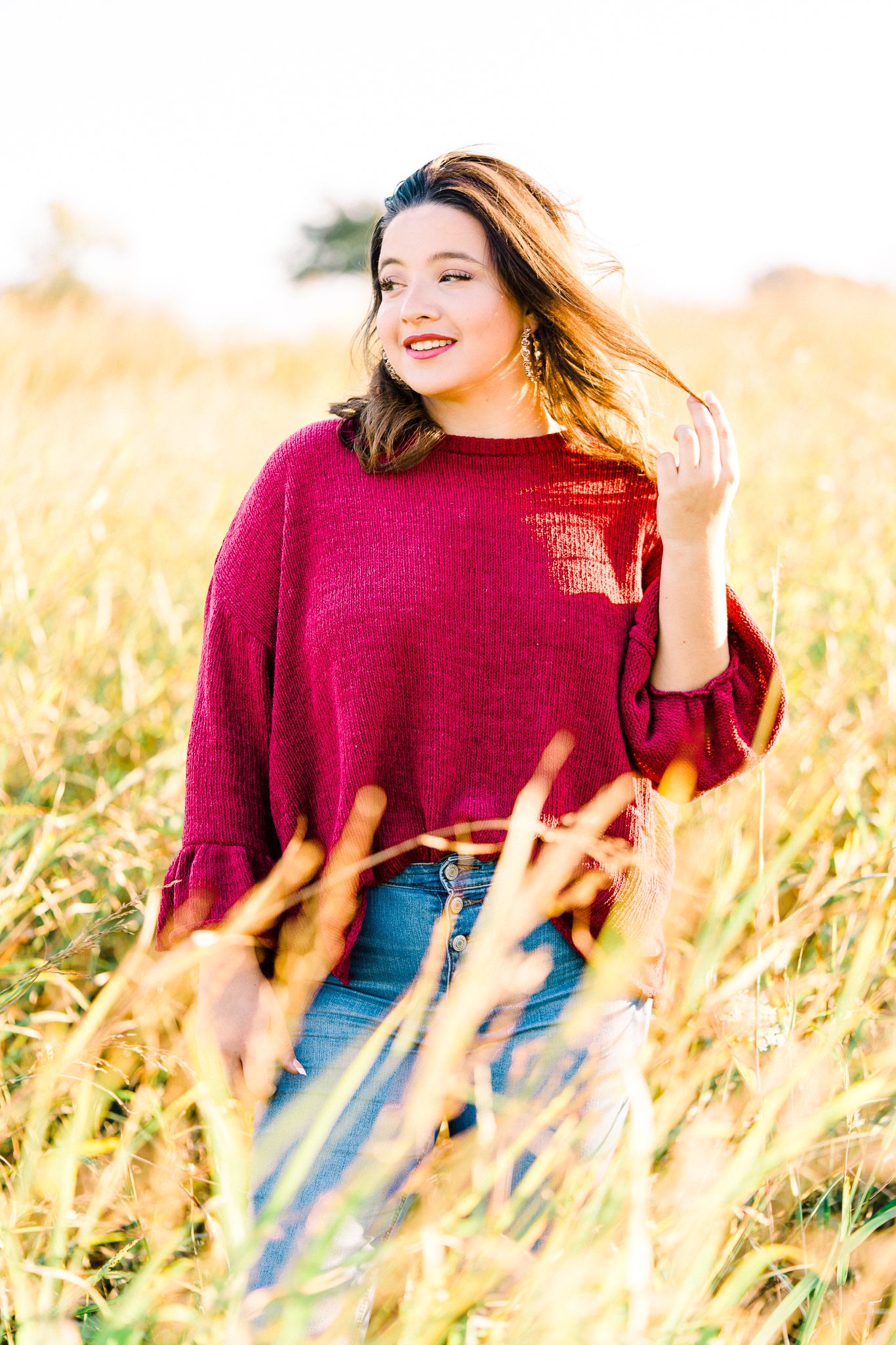 A person in a burgundy sweater and jeans poses playfully in a golden wheat field at sunset.