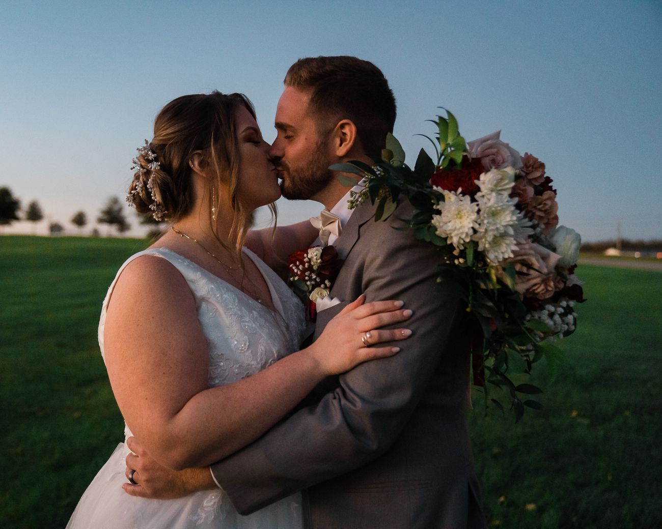 A romantic wedding couple shares an intimate kiss at sunset in a grassy field with blue sky in the background.