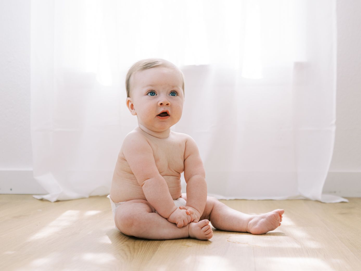 Baby sitting on white floor against bright window background.