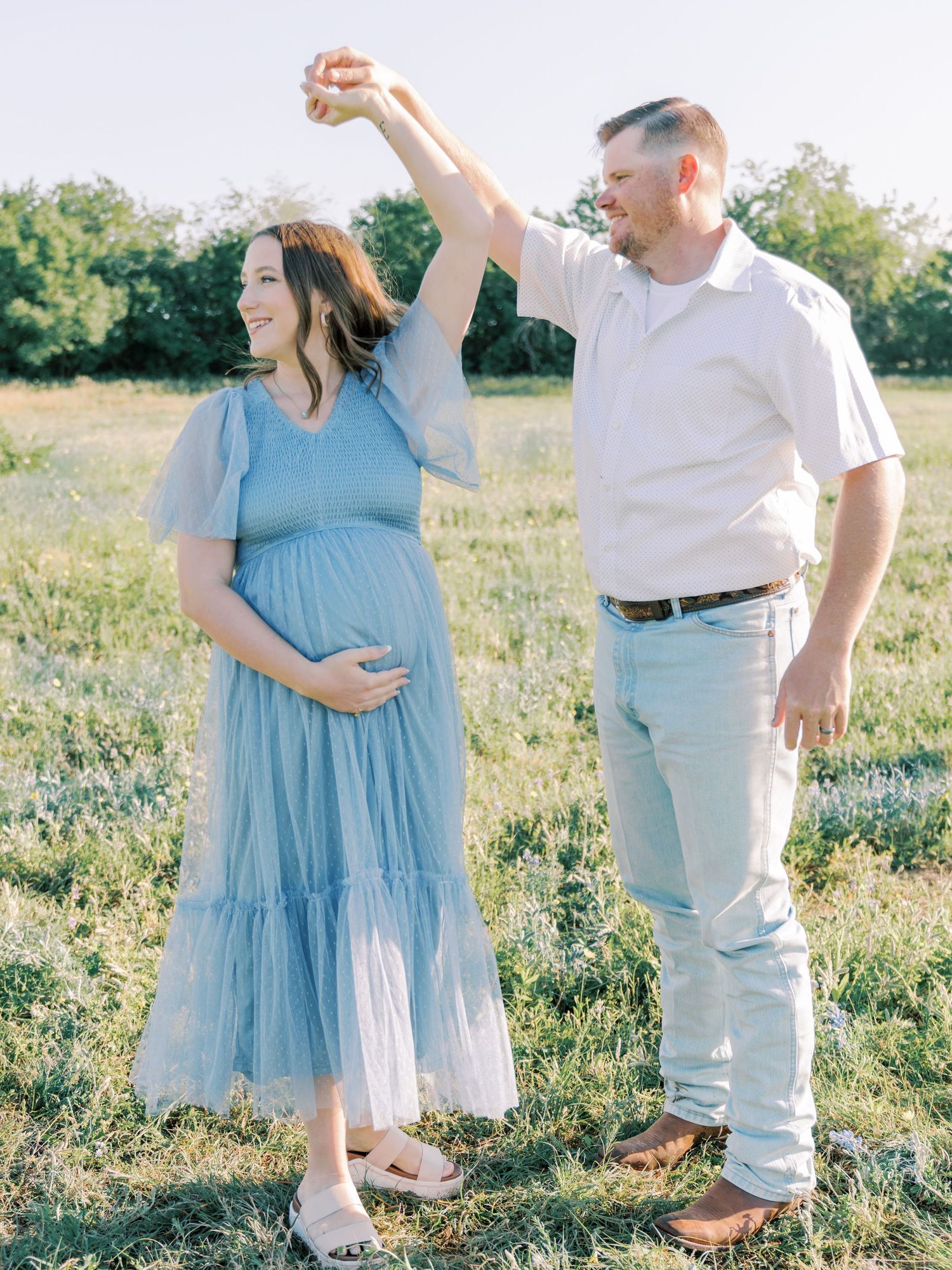 A couple stands together in a sunlit meadow wearing coordinating blue and white outfits during a maternity photoshoot.
