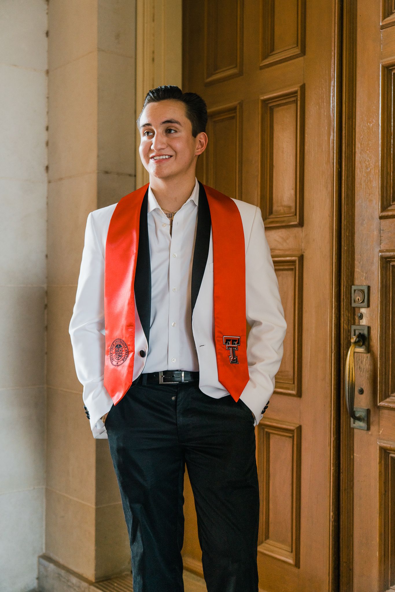 Graduate wearing white dress shirt, black pants and red stole poses against wooden doors in academic hallway.