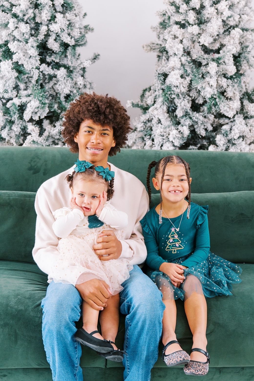 Siblings pose together on an emerald velvet sofa with a frosted Christmas tree backdrop for holiday portraits.