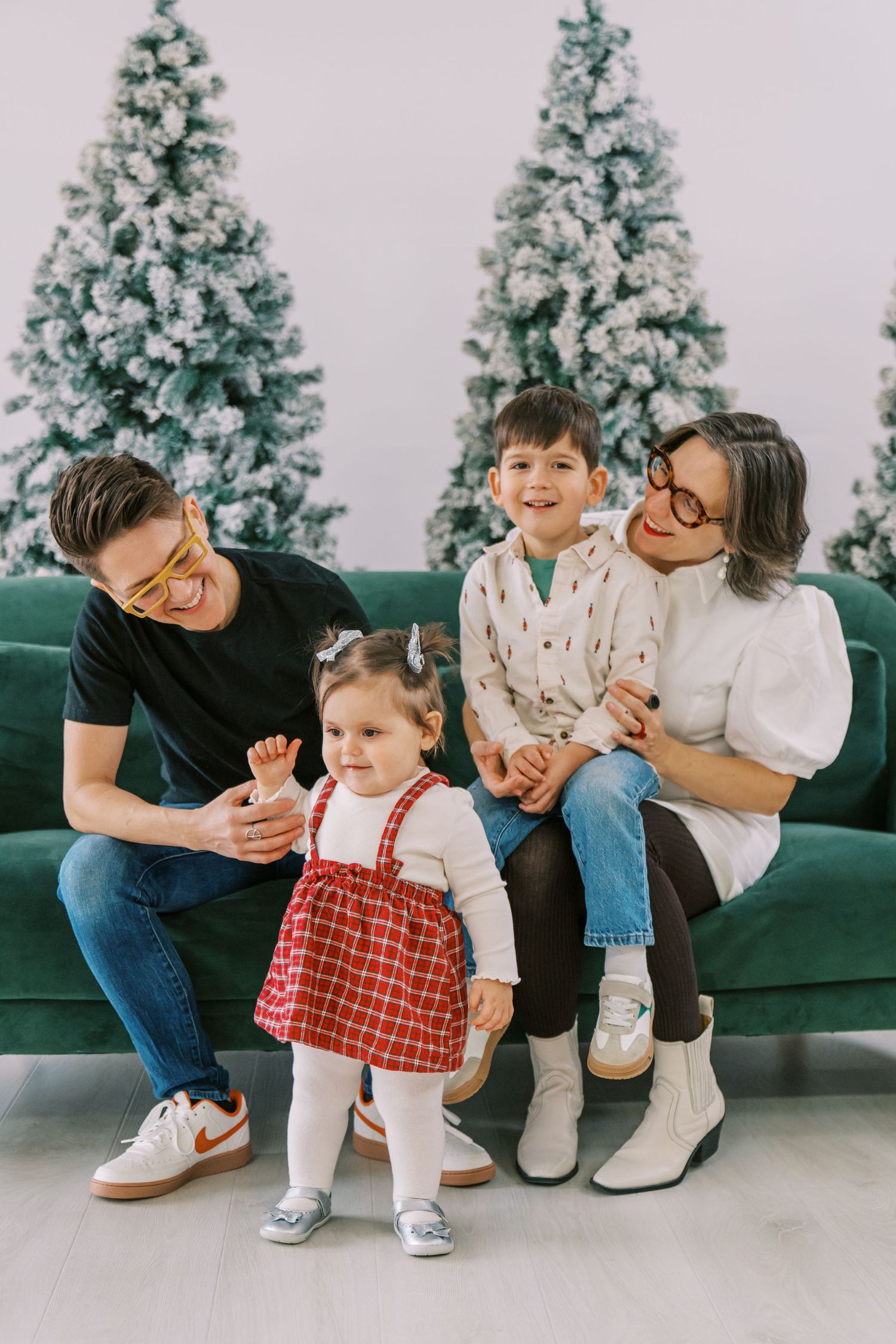Family of four posing on green sofa with snow-flocked Christmas trees, parents with boy and toddler girl in festive outfits.