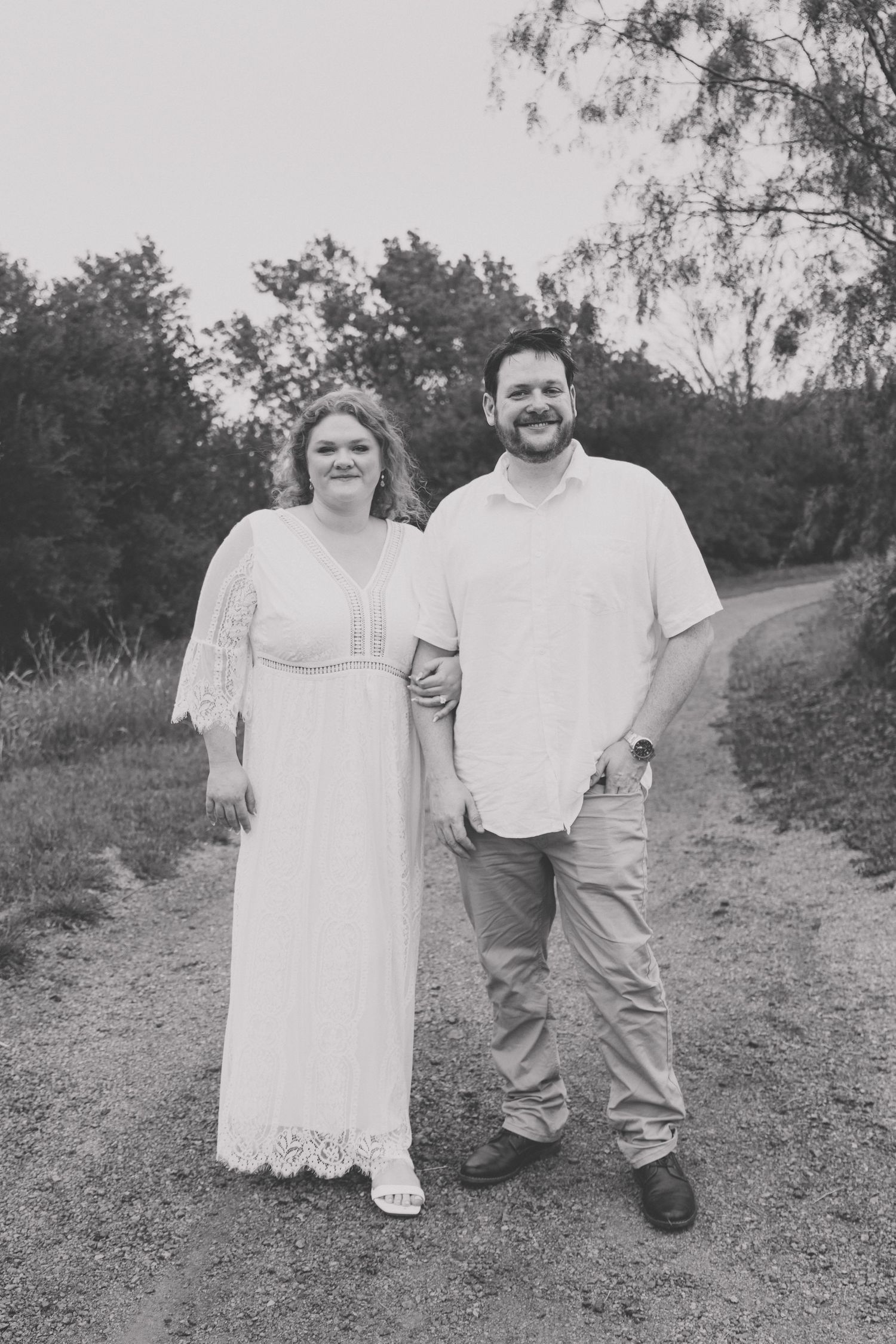 Black and white photo of smiling couple standing on a dirt path surrounded by trees in casual attire.
