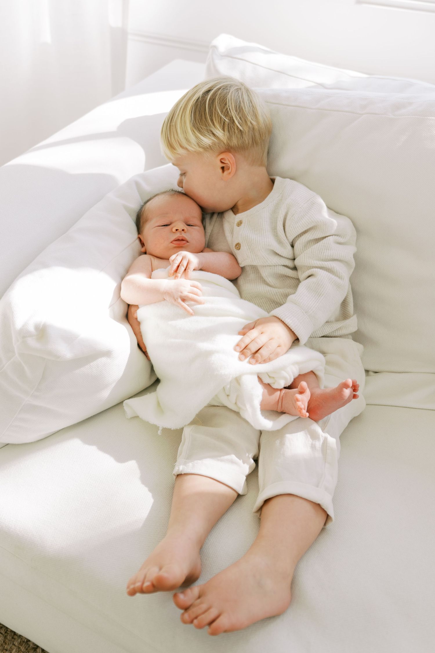 Gentle and intimate series of photos showing a sleeping baby in white bedding being cuddled by a sibling.