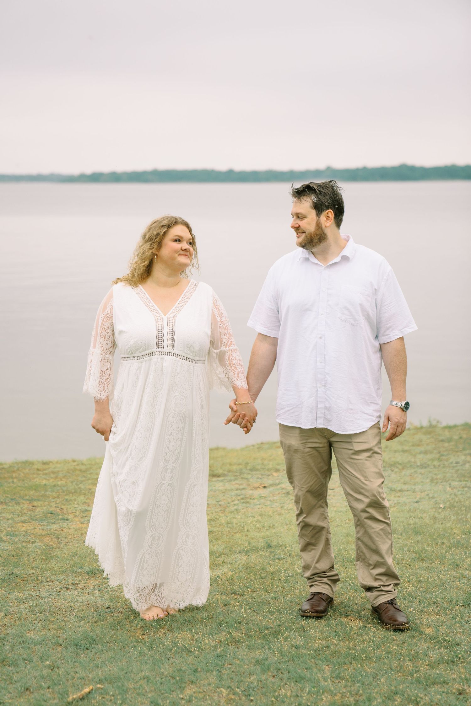 Smiling couple holding hands walking near lakeside, woman in white lace dress and man in white shirt.