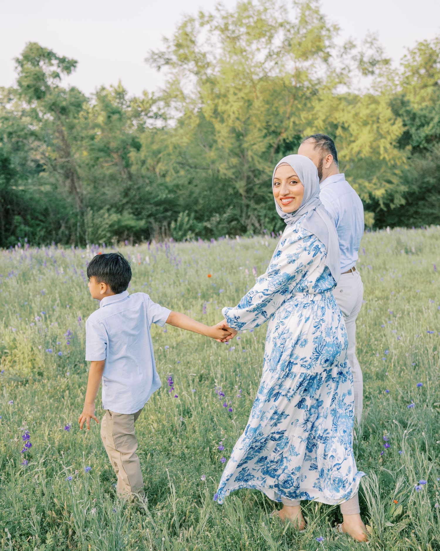 A Muslim family walks through a wildflower meadow, mother in blue floral dress and hijab holding son's hand.