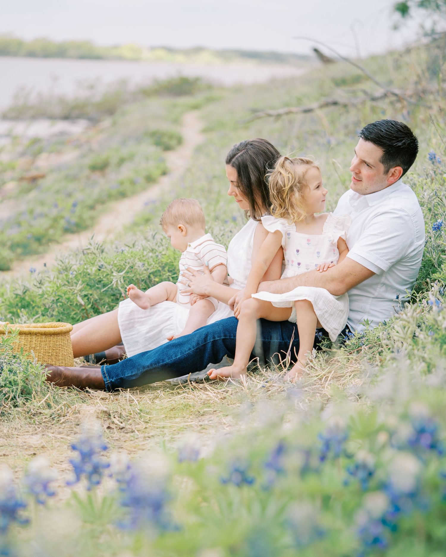 Family of four sitting in a field of bluebonnets near a lake, enjoying a peaceful outdoor picnic together.