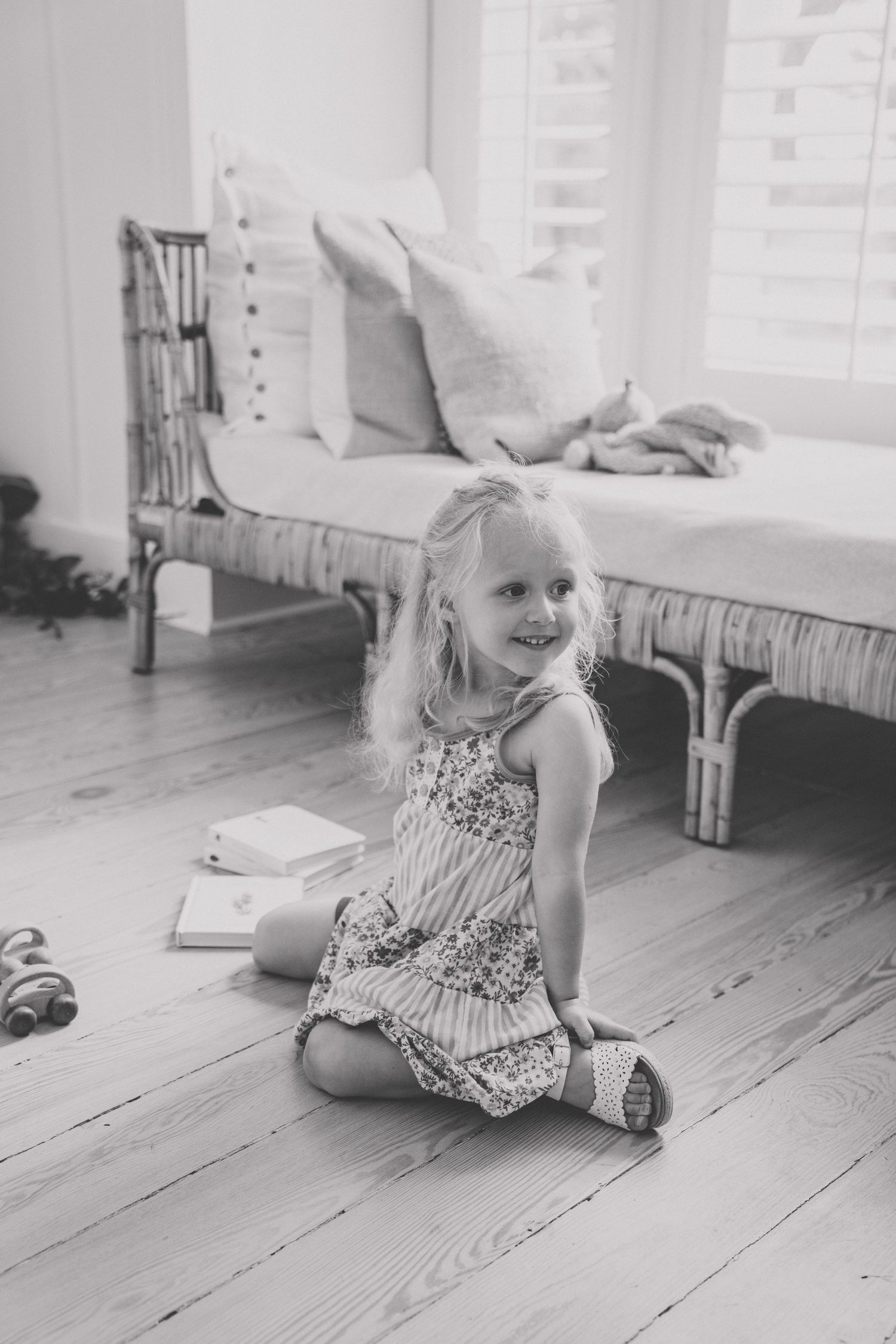 Young blonde girl in floral dress sitting cross-legged on hardwood floor, smiling, with rattan daybed and books nearby.