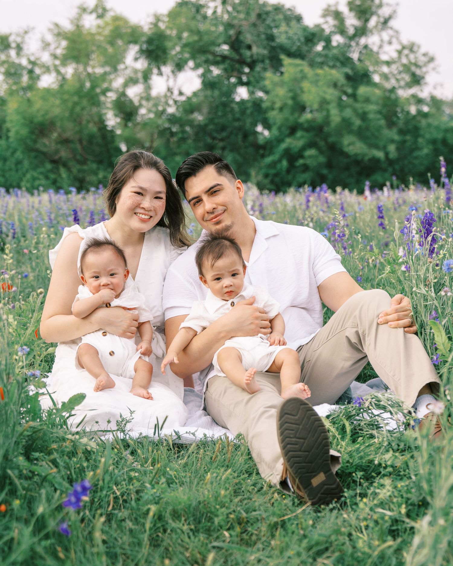 A young family with twin babies sits in a blooming wildflower field, dressed in white and beige tones.