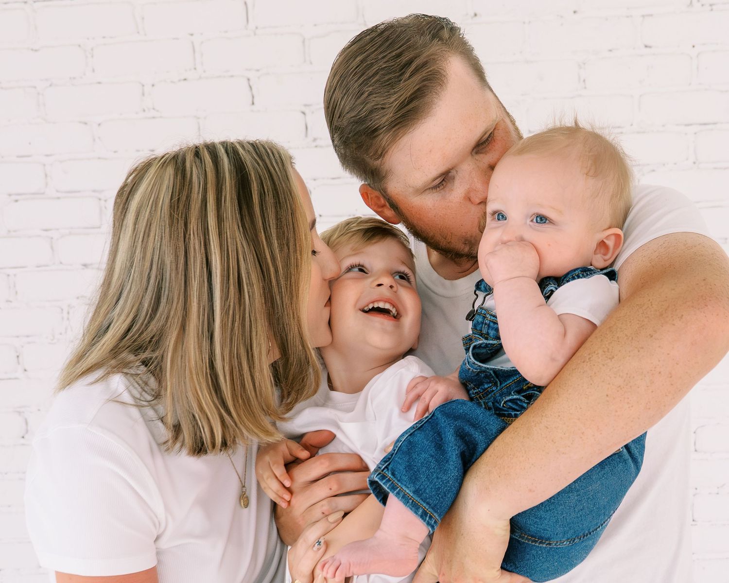 Happy family with parents kissing toddler and baby against white brick wall background.