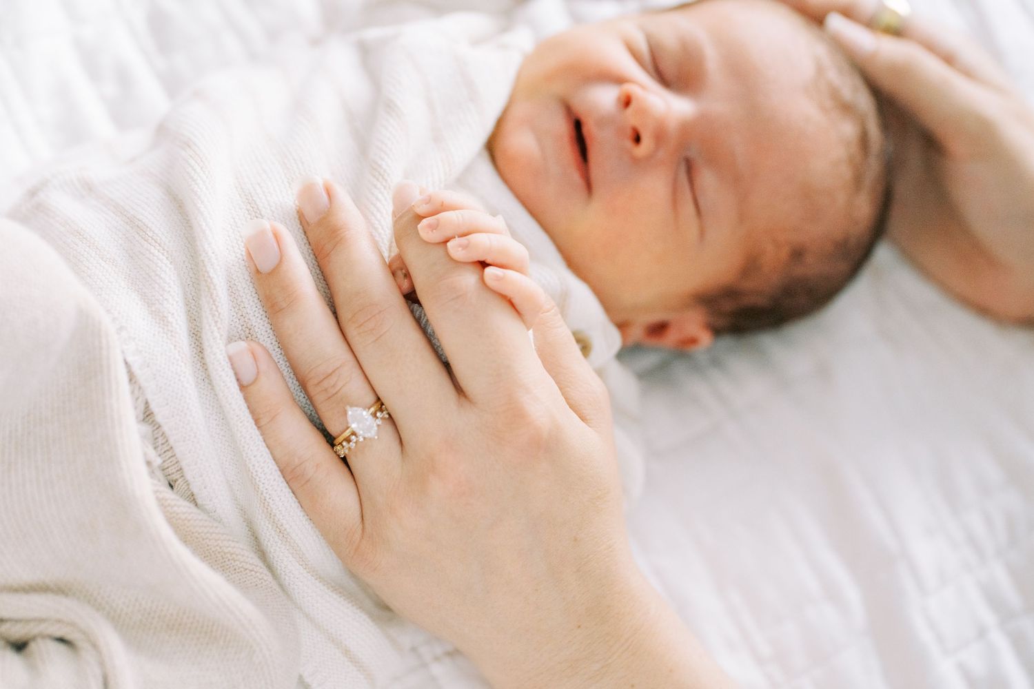 Newborn baby sleeping peacefully on white bedding with hands near face in natural light.