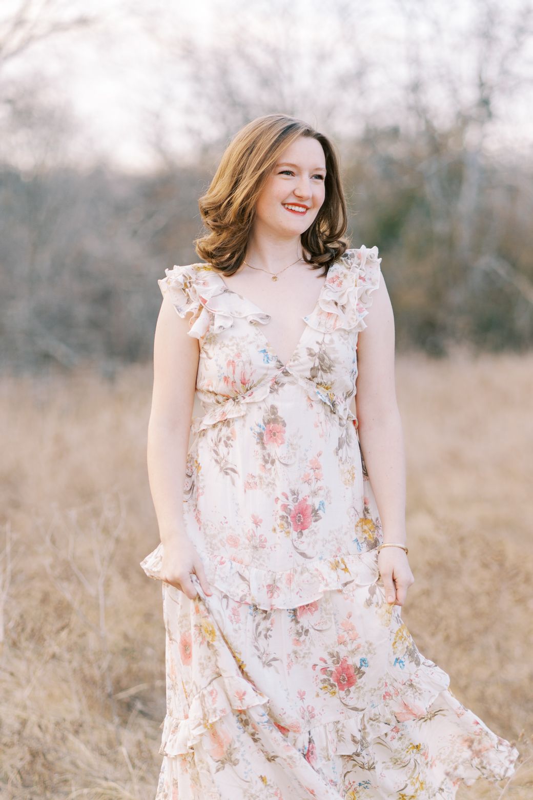 A woman in a flowing floral dress stands in a golden field during sunset with soft dreamy lighting.