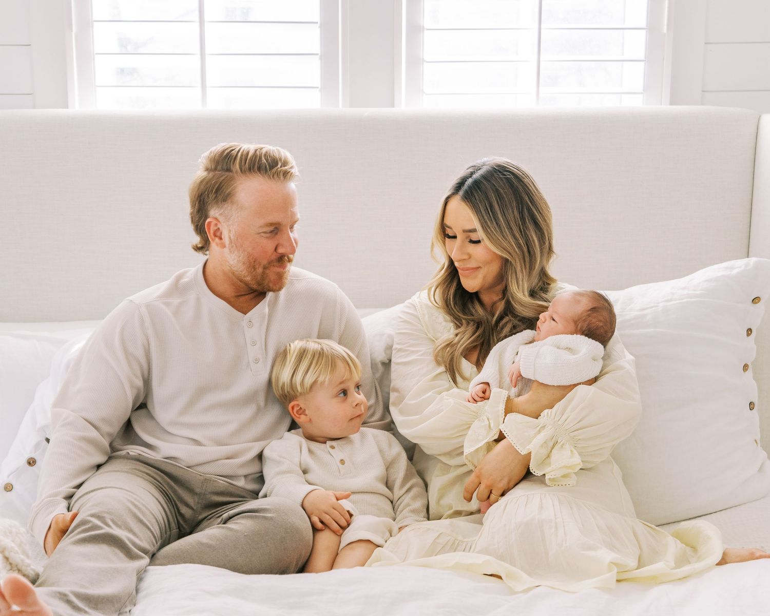 Family of four wearing coordinated neutral outfits sits together on white couch.