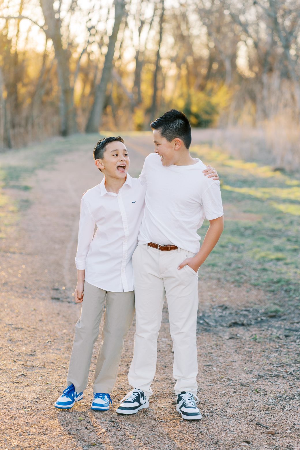 Two young boys in white outfits and blue sneakers share a laugh together on a dirt path in a sunlit forest.