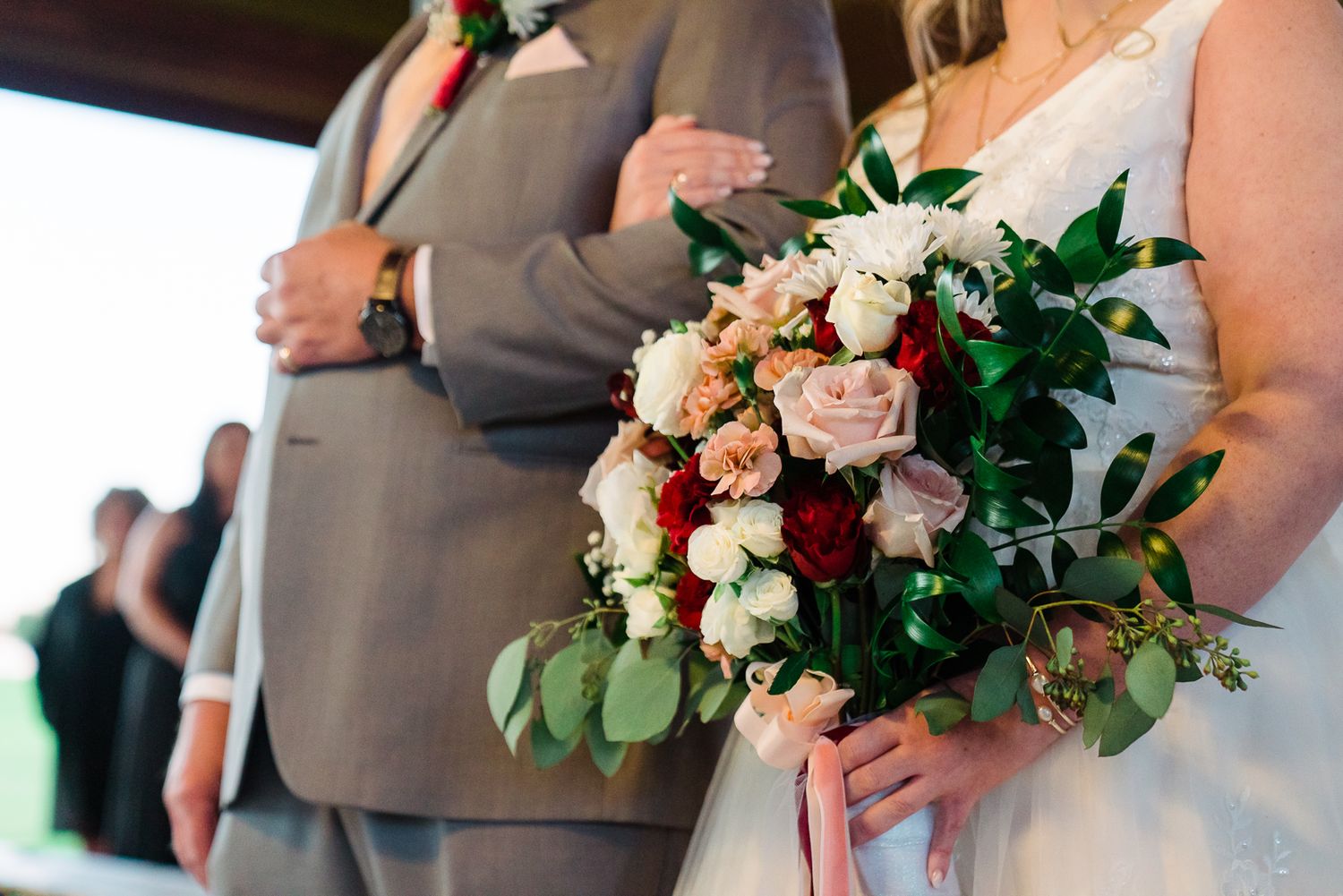 Wedding party members hold elegant bouquets of blush roses and white flowers with greenery against neutral attire.