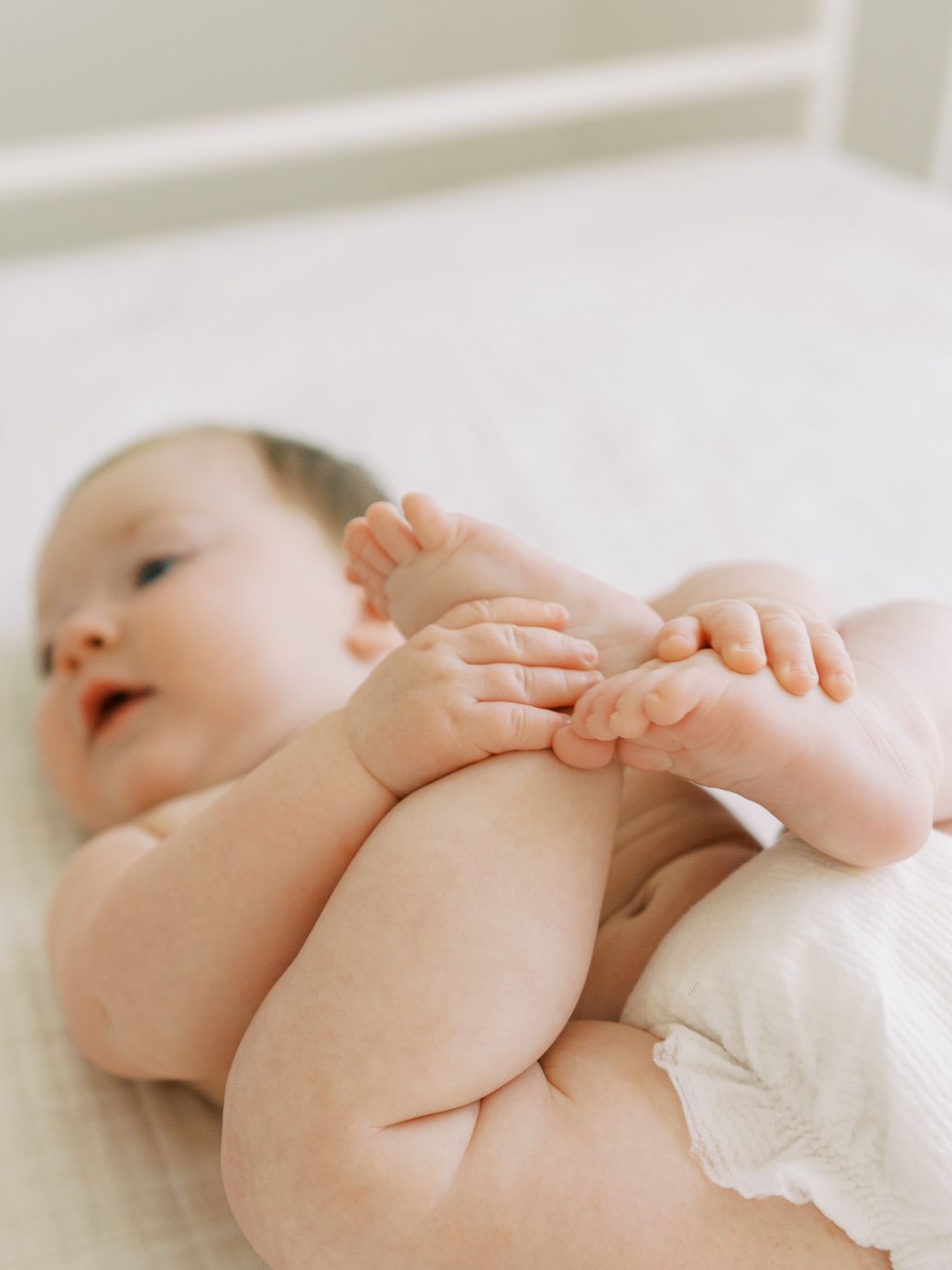Sweet baby lying on white bedding reaching for toes in natural light.