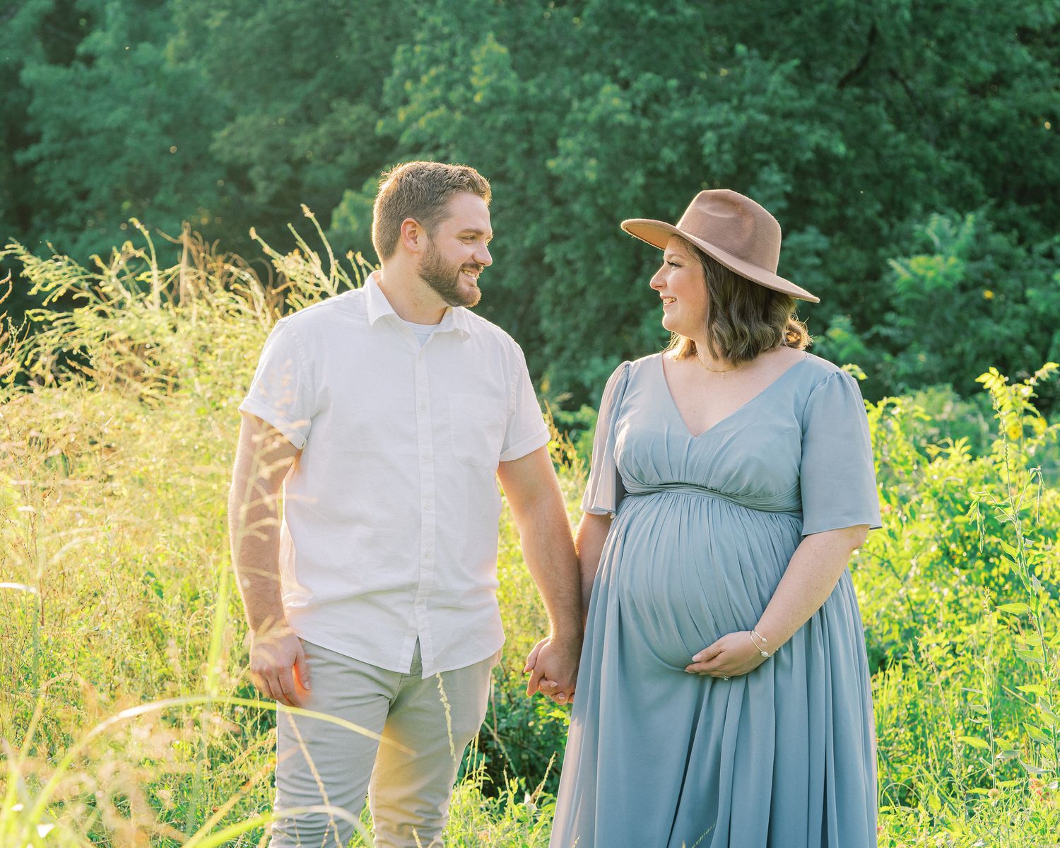 A couple stands together in a sunlit meadow with tall yellow wildflowers and green trees in the background.