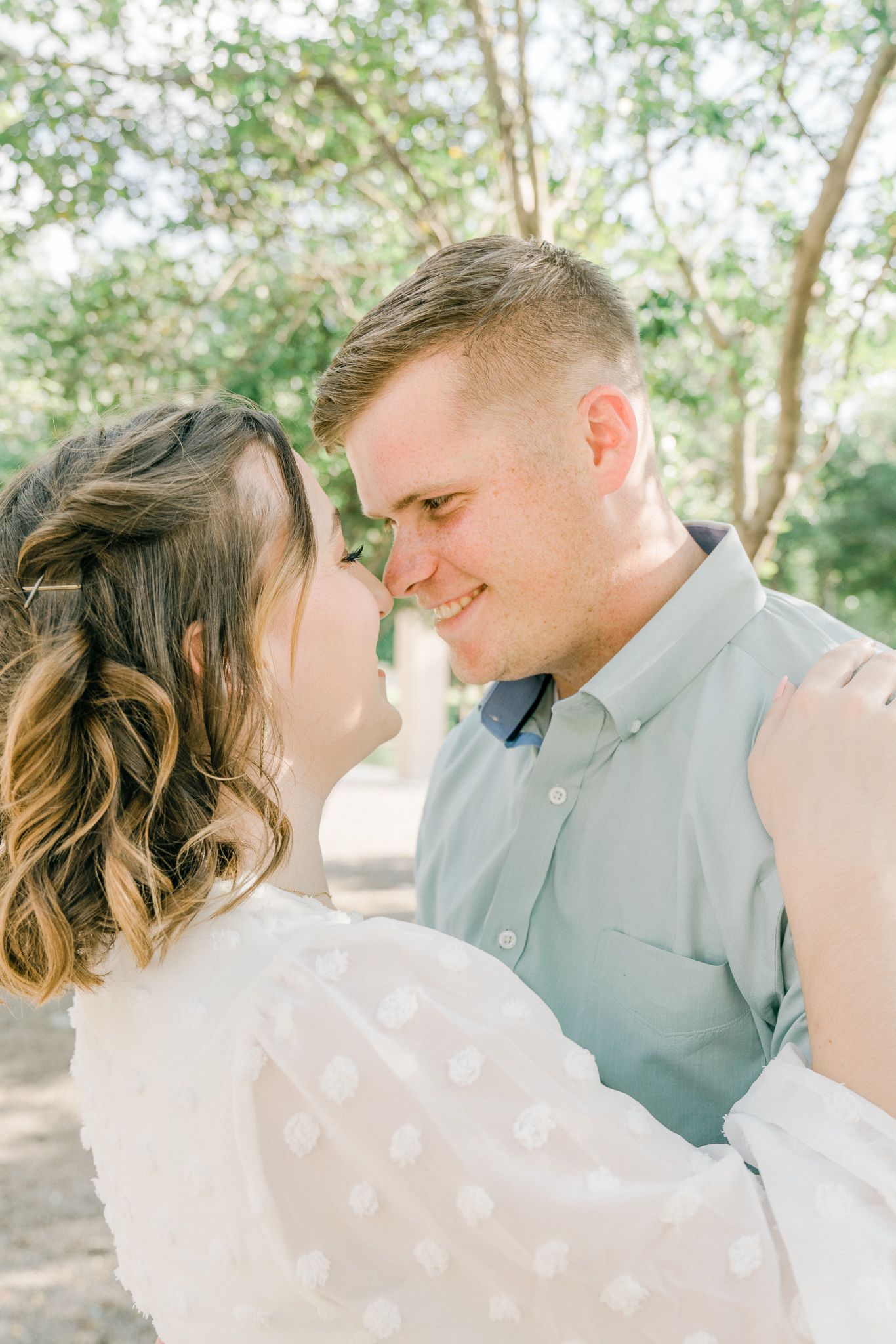 A romantic couple shares intimate moments in a sun-dappled outdoor setting wearing light colored outfits.