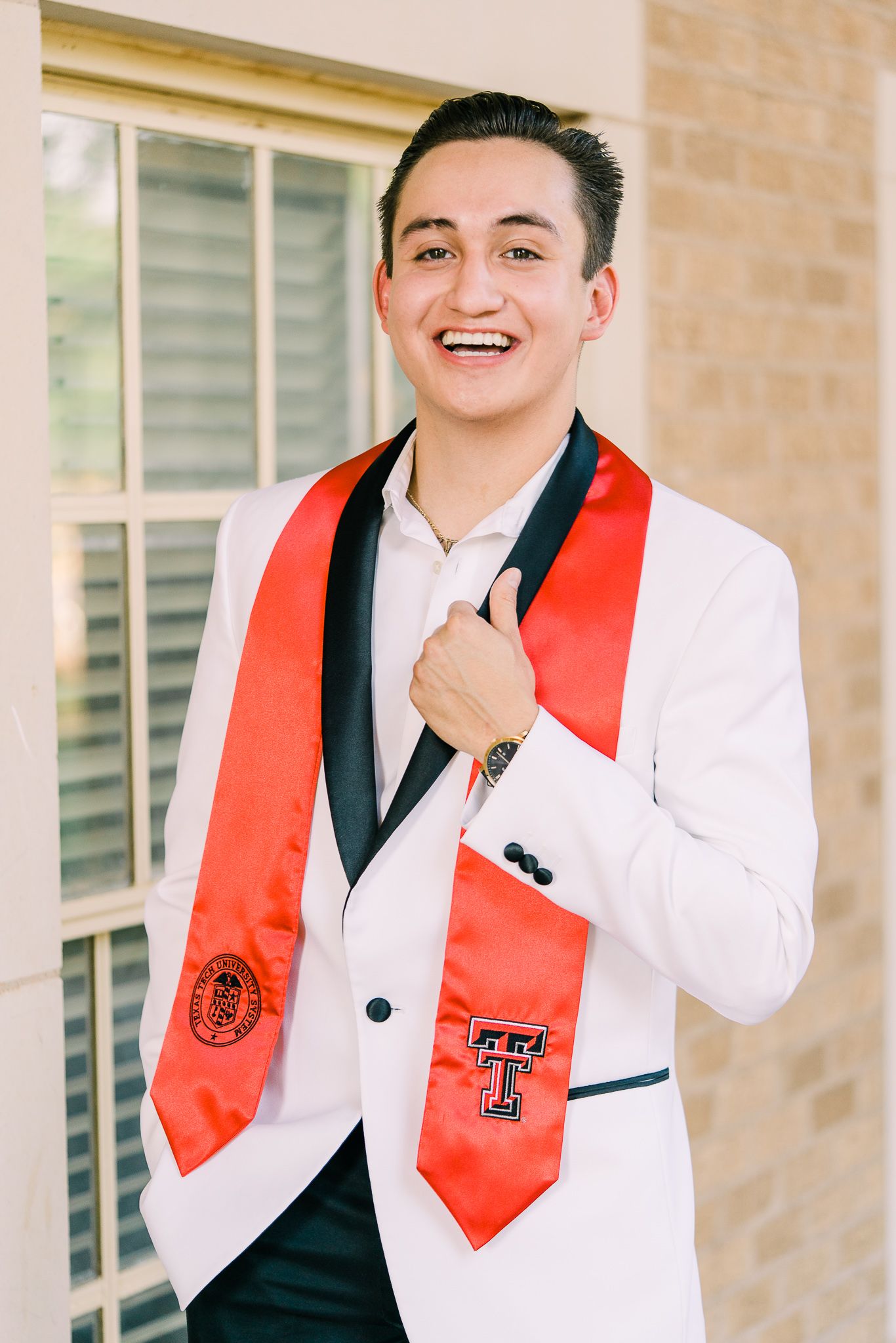 Graduate wearing a white suit jacket and red stole smiles while posing for graduation photos outdoors.