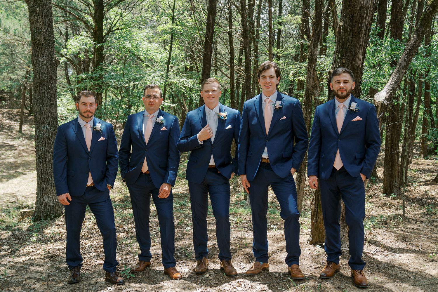 A wedding party wearing navy blue suits poses together in a sunlit forest setting.