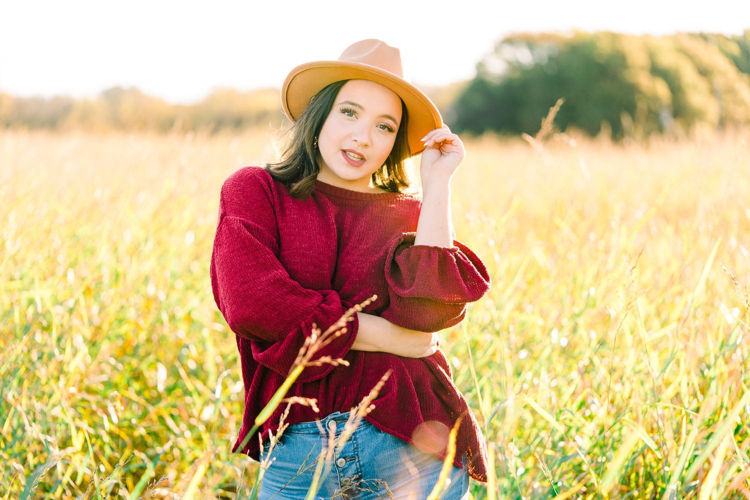 Person in a burgundy sweater and straw hat sits playfully in a golden autumn field during sunset.