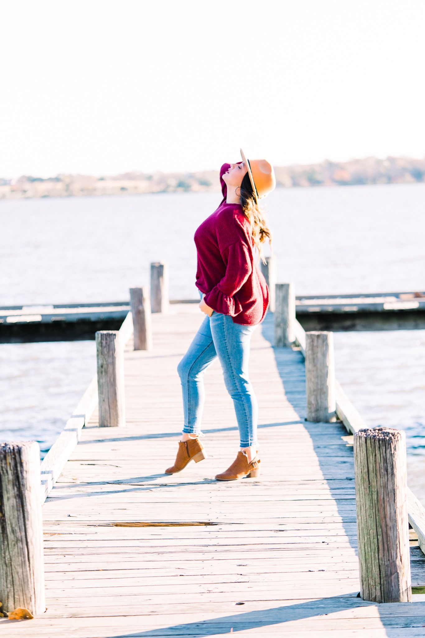 Someone in a burgundy hoodie and jeans stands on a wooden dock by the water on a sunny autumn day.