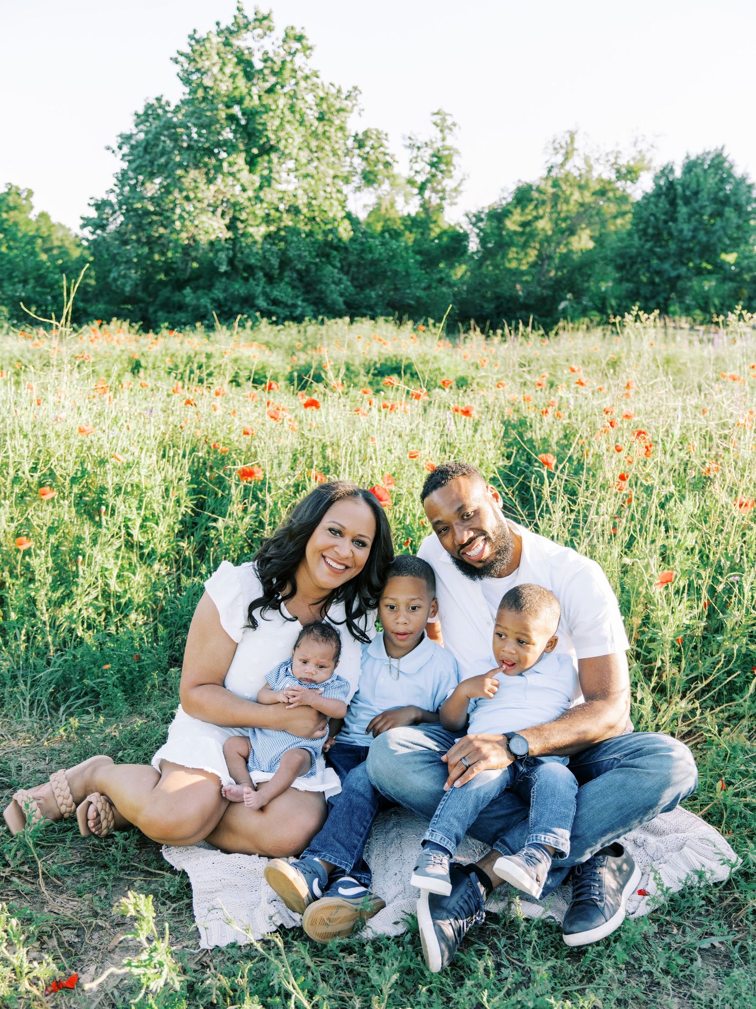 A family sits together in a wildflower meadow during a summer photo session, wearing coordinated white outfits.
