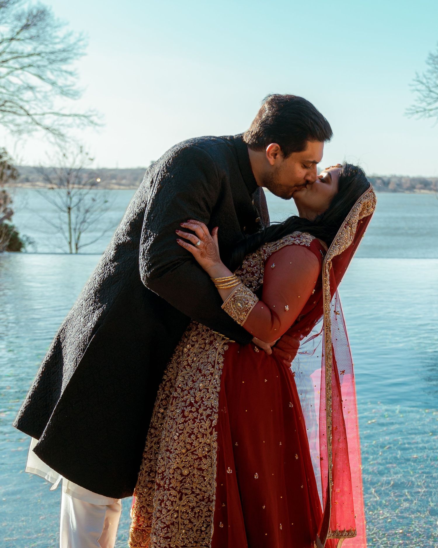 A couple in formal South Asian attire shares intimate moments by a serene lakeside setting.