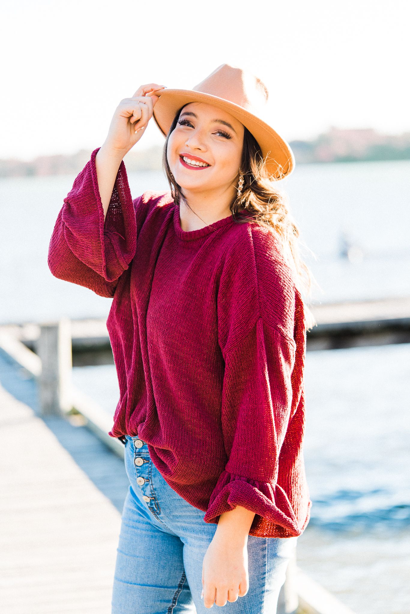 A person in a burgundy sweater and jeans poses playfully by the water on a bright sunny day.