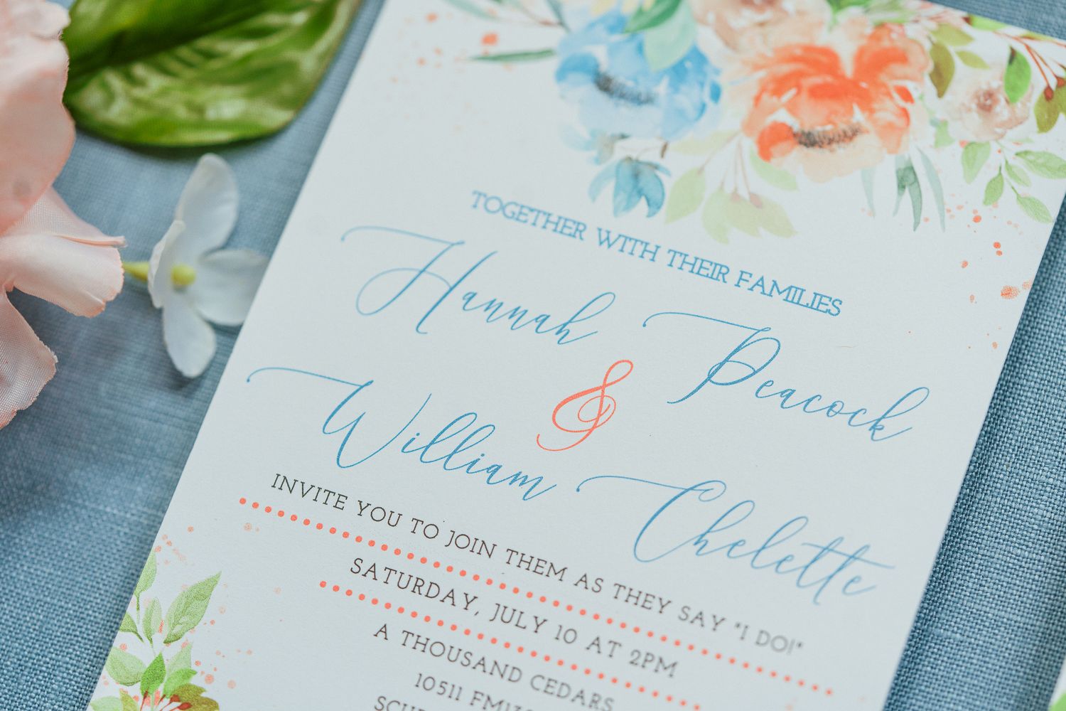 Elegant wedding invitations with blue and coral watercolor floral designs on light blue linen.