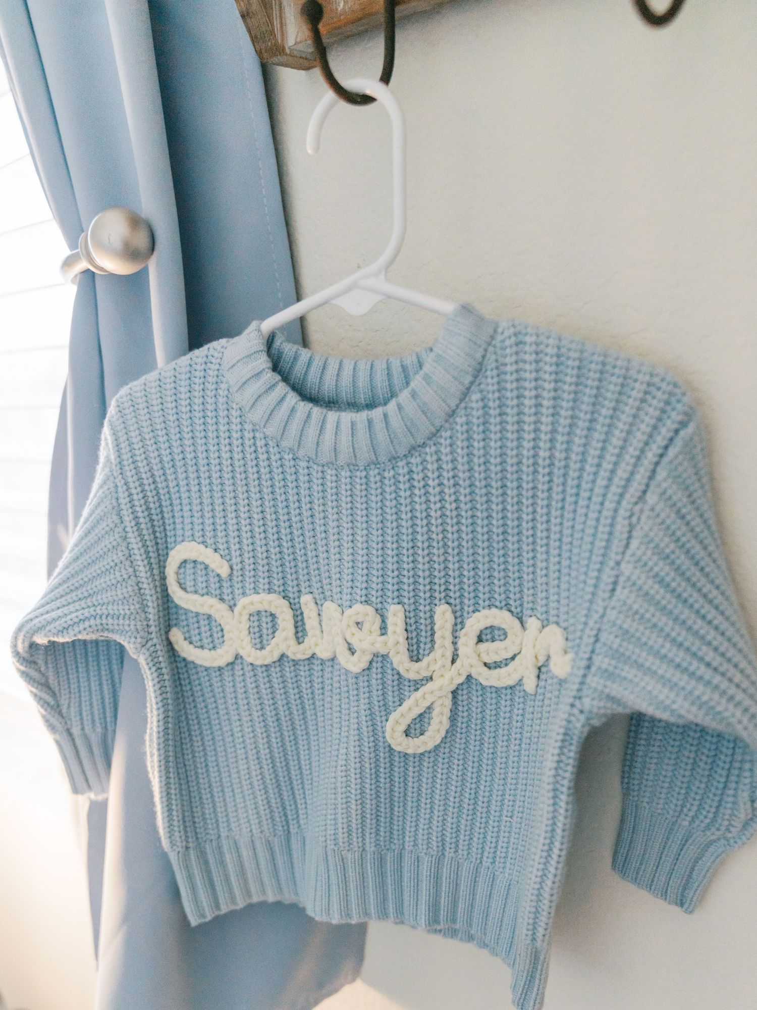 Light blue knit baby sweater with 'Sawyer' embroidered in white hanging on display.
