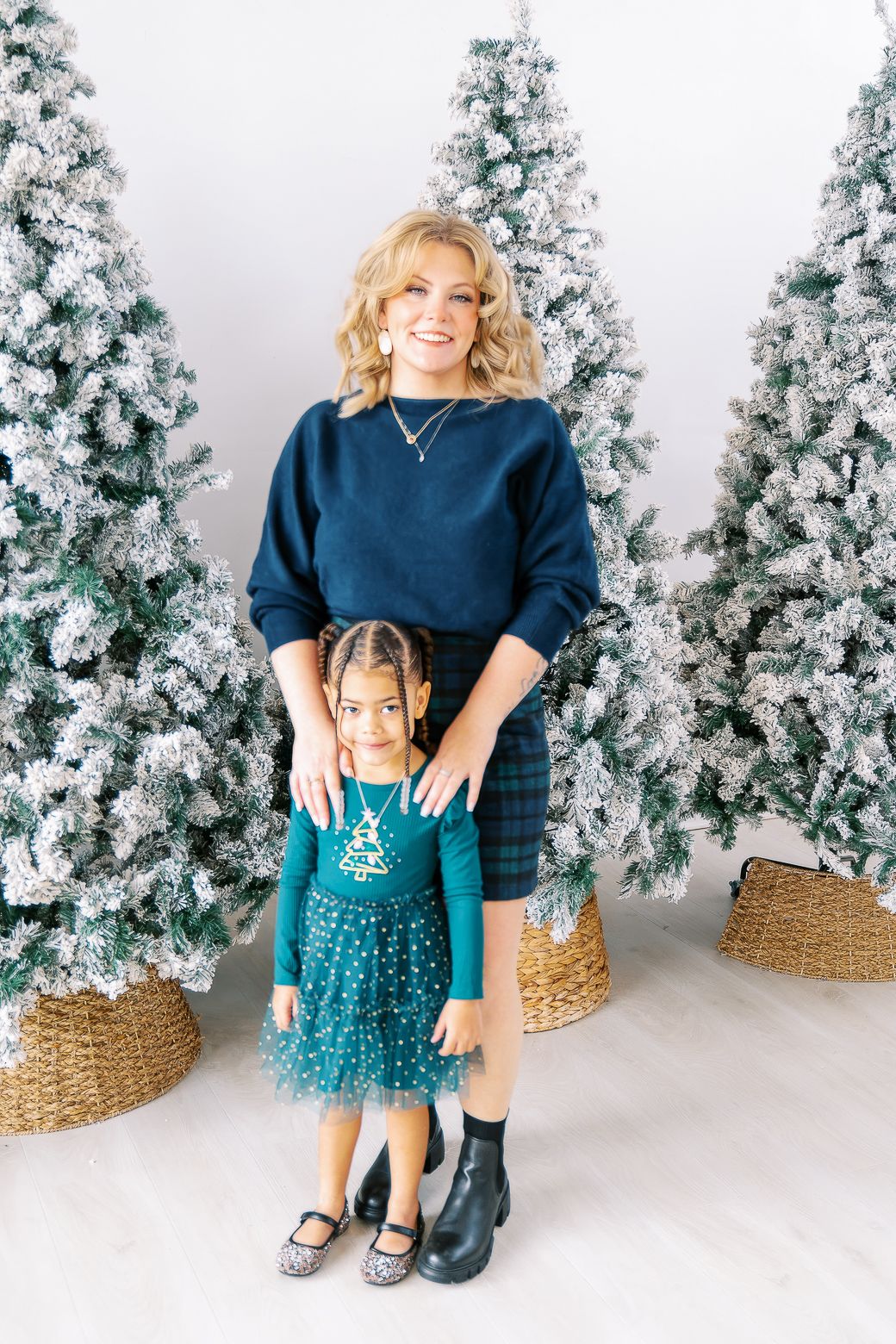 Mother and daughter wearing coordinating navy and teal dresses pose in front of snowy Christmas trees in a studio setting.