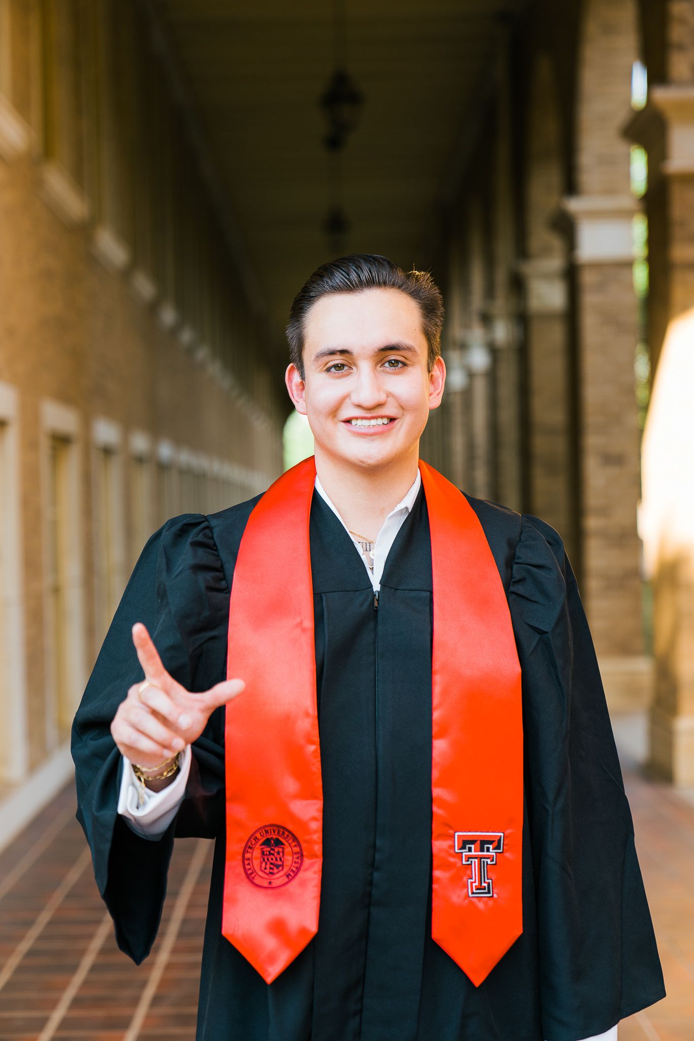 Graduate wearing Texas Tech regalia gives thumbs up while posing in architectural corridor.