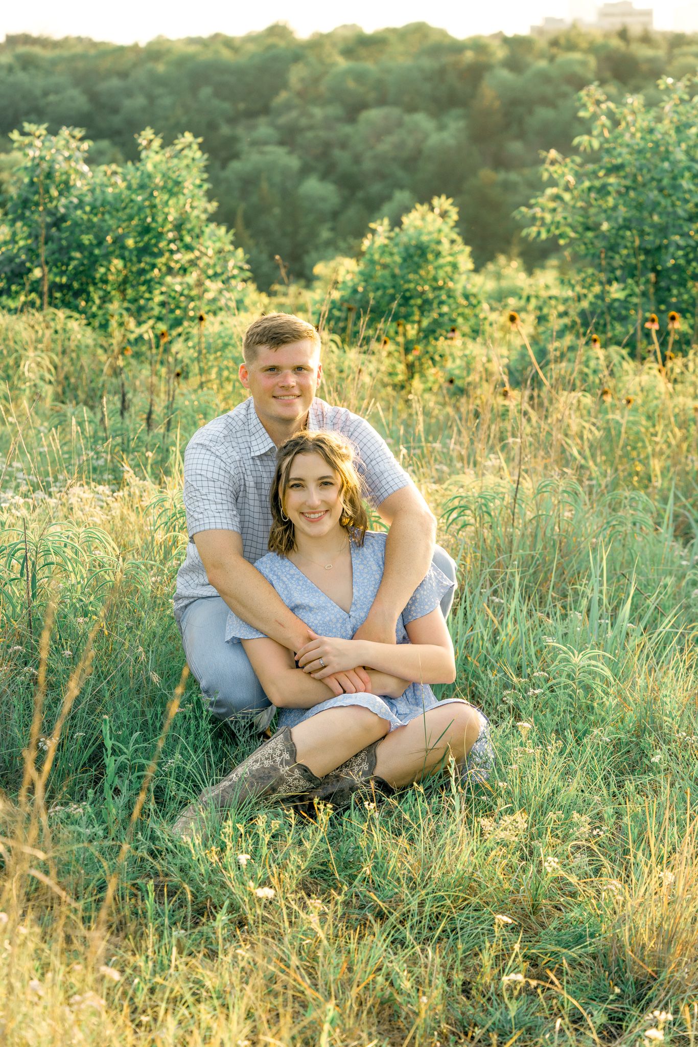 A romantic couple sits together in a sunlit meadow with tall grass and wildflowers during golden hour.